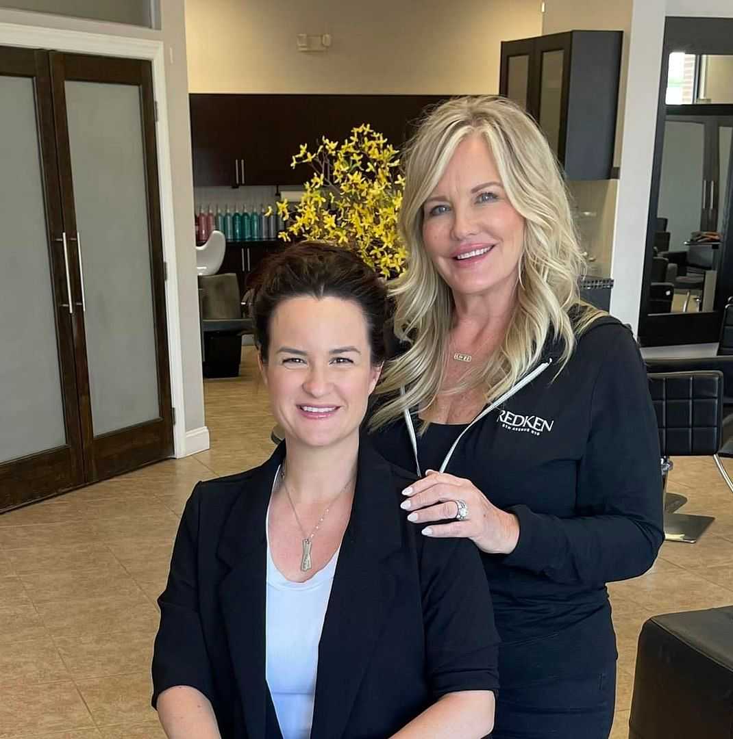 Two women, one seated and the other standing, smile in a salon with modern decor.